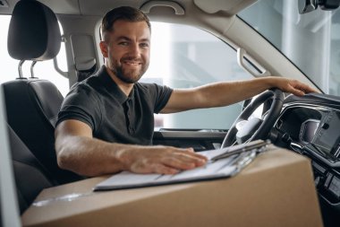 Kameraya bakıyor, gülümsüyor. Teslimatçı ya da yükleyici kutuyla birlikte arabada..