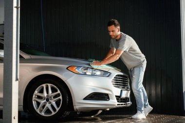 Adam kumaş kullanarak otomobilin yüzeyini cilalıyor. Adam dışarıda arabasıyla bekliyor..