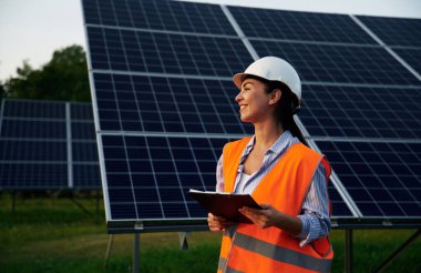 Elinde siyah bir not defteri var. Üniformalı kadın işçi mühendisi güneş panellerinin yakınında..