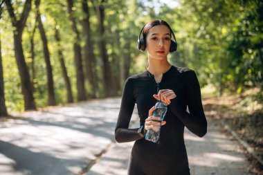 Mola verdim. Kulaklıklar ve suyla ayakta durmak. Genç, sıska kadın dışarıda siyah spor kıyafetleri giyiyor..