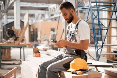Dinleniyor, akıllı telefonla oturuyor. Ahşap depoda sanayi işçisi..