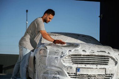 Otomotiv temizleyicisi sabun ve köpüğü yüksek basınçlı çamaşır makinesiyle yıkıyor. Adam dışarıda arabasıyla bekliyor..