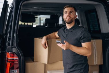 Akıllı telefonu kullanıyordu. Teslimatçı ya da yükleyici elinde bir kutuyla arabanın yanında..