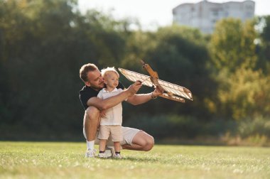Tornavida elimde, tamir ediyorum. Baba ve oğul oyuncak uçakla sahadalar..