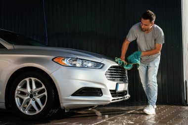 Adam kumaş kullanarak otomobilin yüzeyini cilalıyor. Adam dışarıda arabasıyla bekliyor..