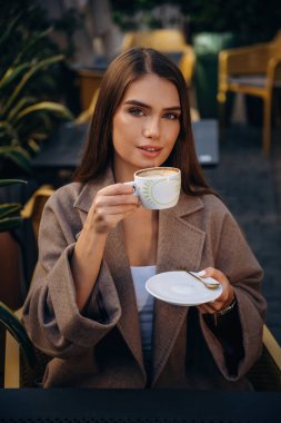 Kameraya bakarak, oturarak. Güzel, genç bir bayan restoranın dışında, masanın yanında..