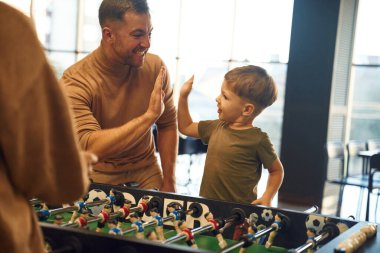 Gülümsüyor, eğleniyor. Ailesi, annesi ve oğlu içeride masa futbolu oynuyorlar..
