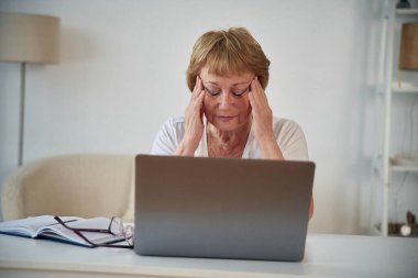Baş ağrısı, dizüstü bilgisayarın yanında oturuyor. Yaşlı yaşlı kadın evde, oturma odasında..