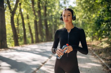 Mola verdim. Kulaklıklar ve suyla ayakta durmak. Genç, sıska kadın dışarıda siyah spor kıyafetleri giyiyor..
