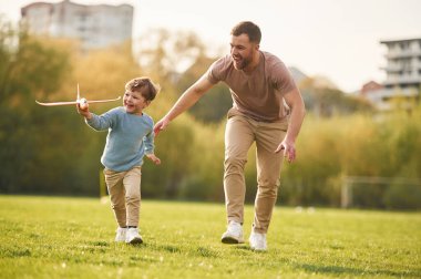 Günlük kıyafetlerle. Oyuncak uçakla koşuyor. Mutlu baba ve oğul yazın sahada eğleniyorlar..