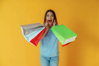 Sürpriz ve beklenen, alışveriş torbalarıyla. Genç bayan sarı arka plana karşı..