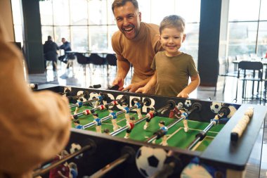 Gülümsüyor, eğleniyor. Ailesi, annesi ve oğlu içeride masa futbolu oynuyorlar..