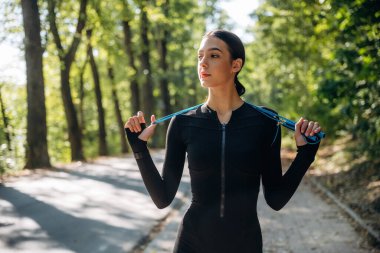 Güneşin altında, mavi atlama ipiyle, ayakta. Genç, sıska kadın dışarıda siyah spor kıyafetleri giyiyor..