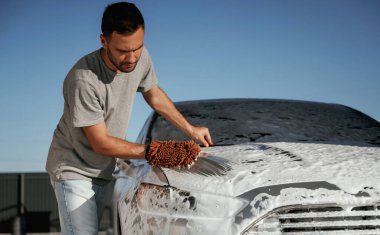 Yüzeyi temizliyor, araba yıkıyor, adam dışarıda arabasıyla bekliyor..