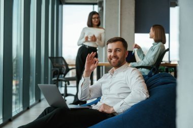 Yakışıklı adam dizüstü bilgisayarla oturuyor. Ofisteki insanlar koltuklarda fasulye torbalarıyla çalışıyorlar..