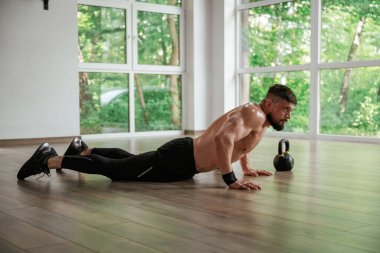 Pencerenin dışındaki yeşil ağaçlar. Güçlü kaslı adam spor salonunda çalışıyor..