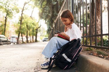 Akıllı telefon elinde, oturuyor. Sırt çantalı kız öğrenci dışarıda..