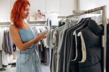 Kızıl saçlı kadın perakende mağazasında kıyafet seçiyor..