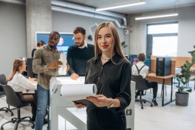 Kadın meslektaşlarının önünde duruyor. Ofiste 3D yazıcının yanında insanlar var..