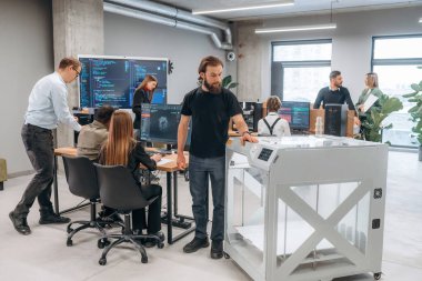 Duvarında kod olan büyük ekran. Ofiste 3D yazıcının yanında insanlar var..