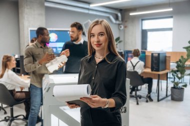 Kadın meslektaşlarının önünde duruyor. Ofiste 3D yazıcının yanında insanlar var..