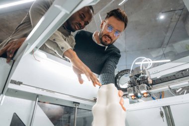 Yazdırılmış nesnenin odaklanmış görünümü. Ofiste 3D yazıcının yanında insanlar var..