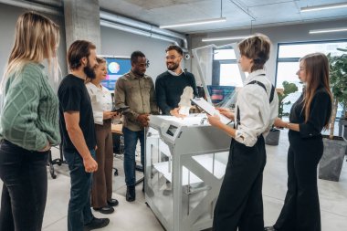 Toplanıyor, basılı ürünlere bakıyor. Ofiste 3D yazıcının yanında insanlar var..