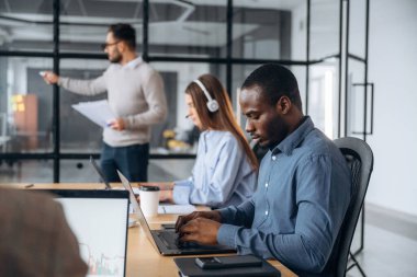 Odaklanmış, dizüstü bilgisayarın yanında oturuyor. Profesyonel işçiler modern ofiste birlikte çalışıyorlar..