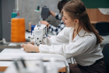 Yan taraftan bak, otur. İnsanlar içeride dikiş makinelerinin yanında çalışıyor..