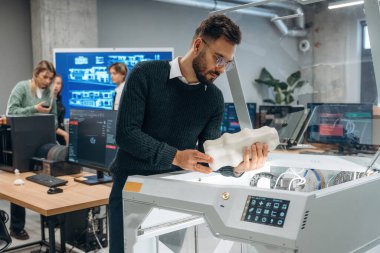 Beyaz baskılı nesneler ellerinde. Ofiste 3D yazıcının yanında insanlar var..