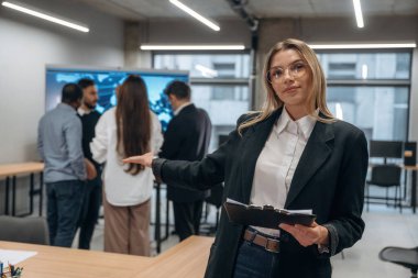 Gözlüklü kadın elinde not defteriyle ayakta duruyor. Bir grup ofis çalışanı içeride birlikte..