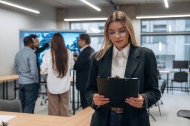 Gözlüklü kadın elinde not defteriyle ayakta duruyor. Bir grup ofis çalışanı içeride birlikte..