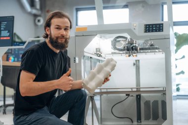 Oturmak ve basılı ürünleri tutmak. Adam 3D yazıcıyla ofiste..