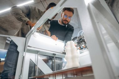 Yazdırılmış nesnenin odaklanmış görünümü. Ofiste 3D yazıcının yanında insanlar var..