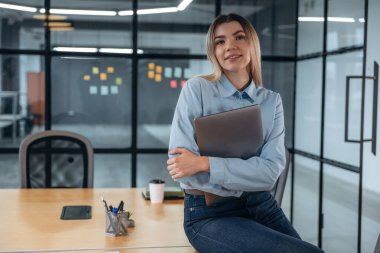 Poz vermek, masada oturmak, dizüstü bilgisayarı tutmak. Kadınlar modern ofiste, resmi kıyafetler giyiyorlar..