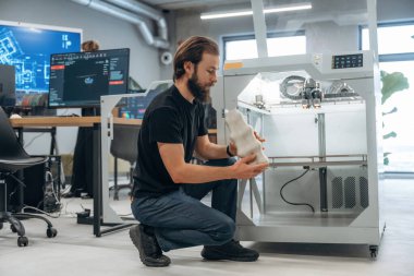 Oturmak ve basılı ürünleri tutmak. Adam 3D yazıcıyla ofiste..