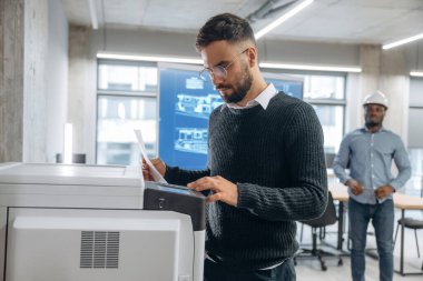 İki ofis çalışanı ayakta. Yazıcının yanında belgeleri olan bir adam var..