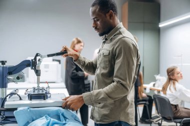 Siyah adam masanın yanında duruyor. İnsanlar içeride dikiş makinelerinin yanında çalışıyor..