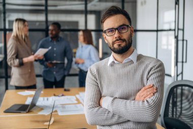 Adam kollarını kavuşturmuş duruyor. Profesyonel işçiler modern ofiste birlikte çalışıyorlar..
