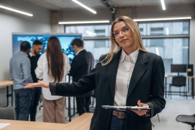 Gözlüklü kadın elinde not defteriyle ayakta duruyor. Bir grup ofis çalışanı içeride birlikte..