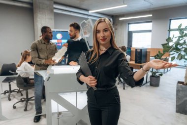 Genç bayan meslektaşlarının önünde duruyor. Ofiste 3D yazıcının yanında insanlar var..