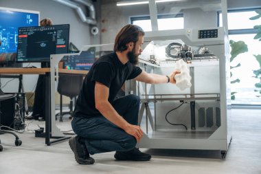 Oturmak ve basılı ürünleri tutmak. Adam 3D yazıcıyla ofiste..