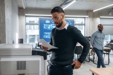 İki ofis çalışanı ayakta. Yazıcının yanında belgeleri olan bir adam var..