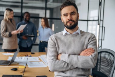 Adam kollarını kavuşturmuş duruyor. Profesyonel işçiler modern ofiste birlikte çalışıyorlar..