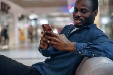 Oturmak ve akıllı telefon tutmak. Siyah adam süpermarketin holünde..