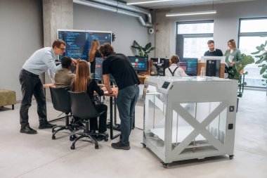 Duvarında kod olan büyük ekran. Ofiste 3D yazıcının yanında insanlar var..