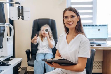 Elinde not defteriyle. Göz doktoru kliniğinde doktorla küçük bir kız..