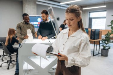 Elinde not defteri var, kadın ayakta. Ofiste 3D yazıcının yanında insanlar var..