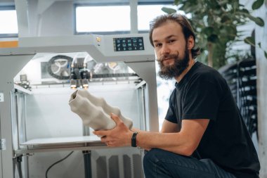 Oturmak ve basılı ürünleri tutmak. Adam 3D yazıcıyla ofiste..