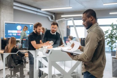 Afrikalı Amerikalı bir adam meslektaşlarının önünde duruyor. Ofiste 3D yazıcının yanında insanlar var..